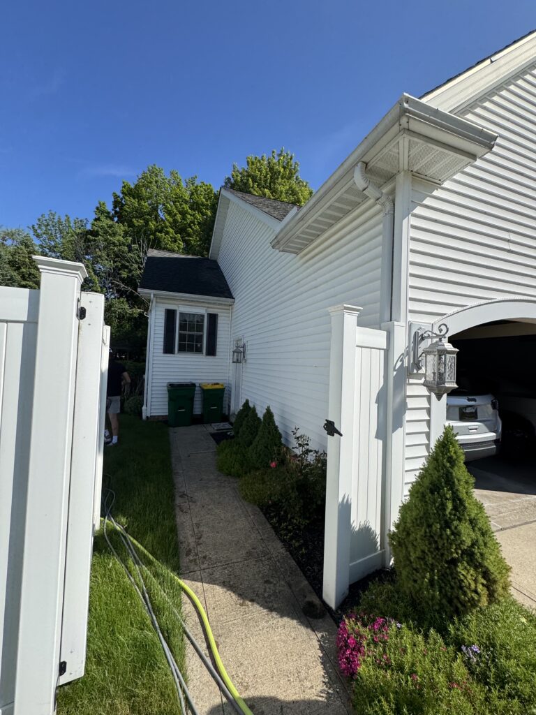 Side view of a white vinyl-siding house with a garage, a concrete path, a nearby green garden hose, and green shrubbery under a clear blue sky.
