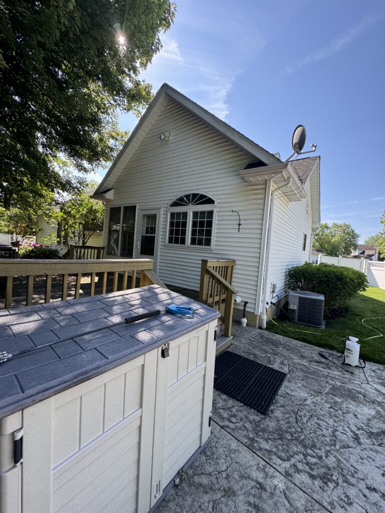 White suburban house with a gabled roof, satellite dish on the side, and a wooden deck in the yard; foreground shows a white storage shed.