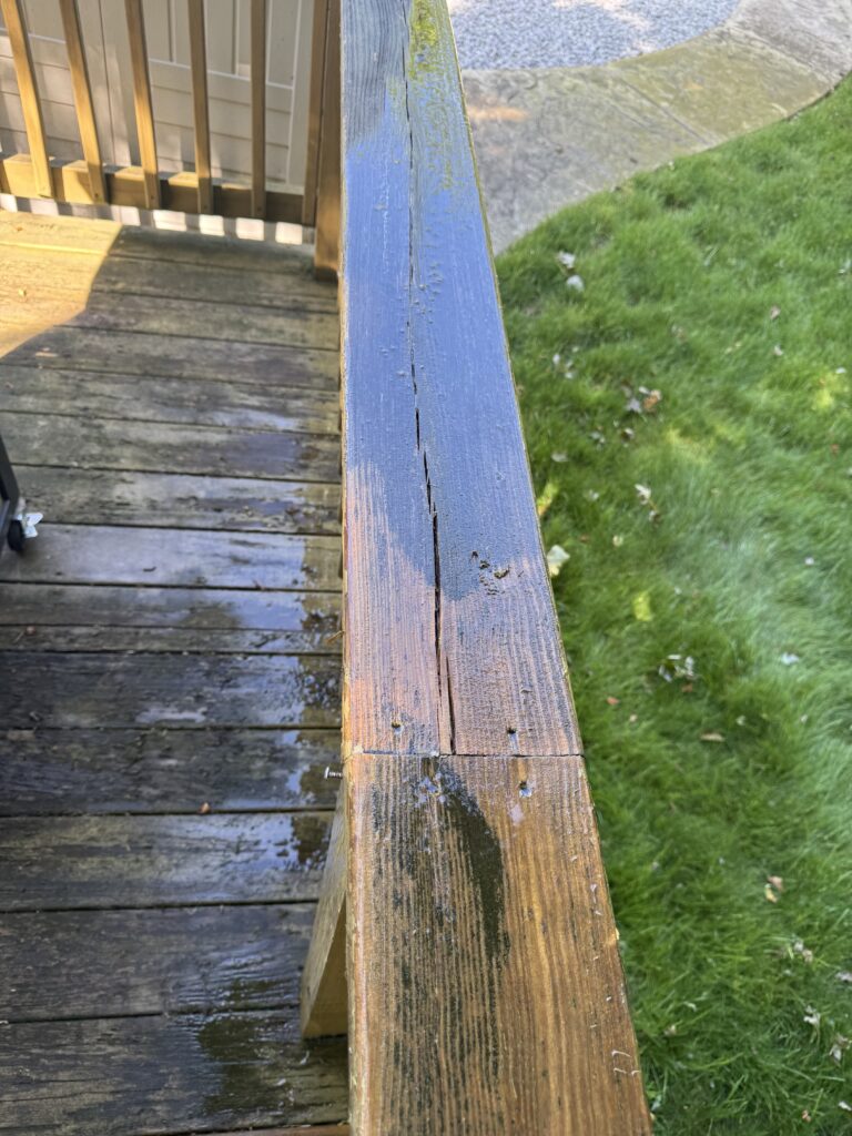 Close-up of a weathered blue wooden railing on a deck, showing peeling paint and a deep crack, with green lawn in the background.