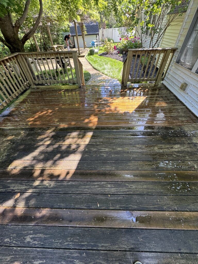 Rain-wet wooden deck with reflections, leading to a sunny backyard garden with grass, flowers, and a person near a small shed in the background.