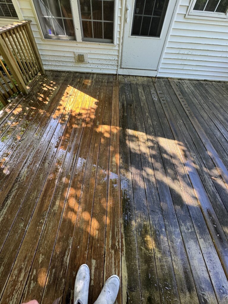 Weathered wooden deck with wet, stained boards in sunlight, next to a white siding house with a glass door and window that show the before and after cleaning of Wiley's Pressure Washing Services