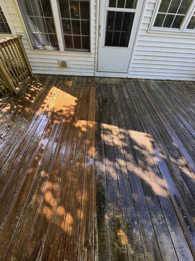 Weathered wooden deck with wet, stained boards in sunlight, next to a white siding house with a glass door and window(s).