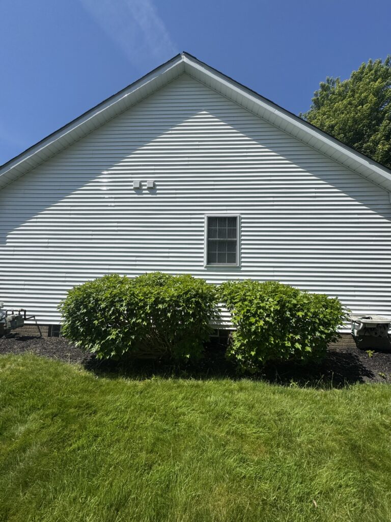 White vinyl-sided house with a prominent triangular gable, a single window, and neatly trimmed shrubs in front under a blue sky.