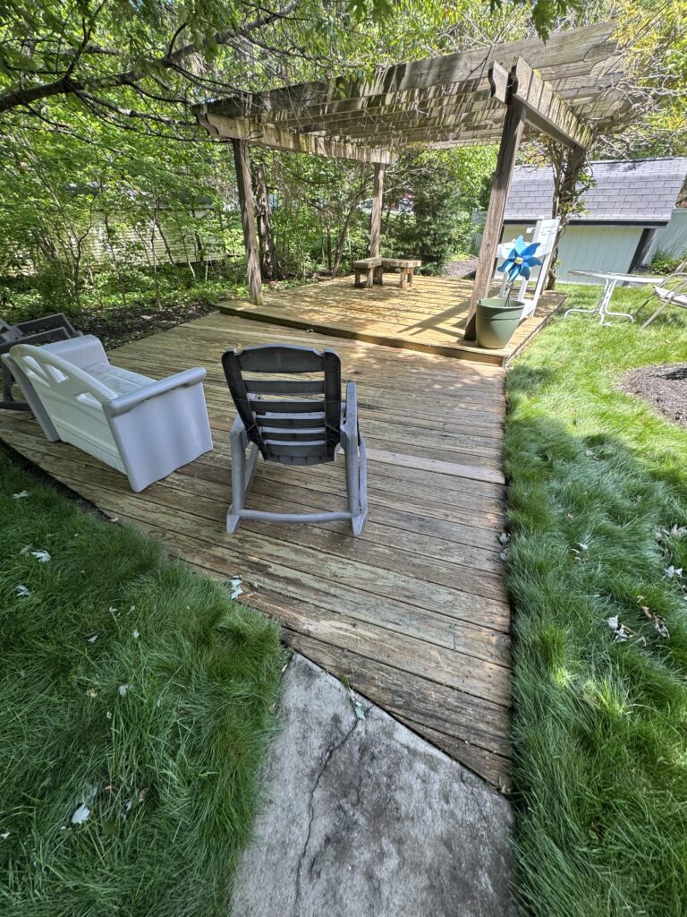 Wooden backyard deck with a rustic pergola, plastic chairs, and a small bench among green plants and trees.