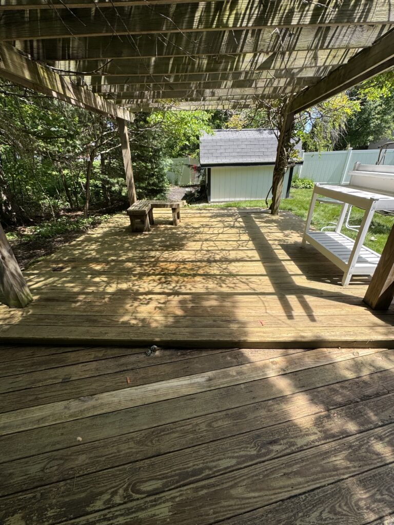 Wooden deck under a vine-covered pergola, small rustic benches, with a shed and green yard beyond a white fence in bright daylight.