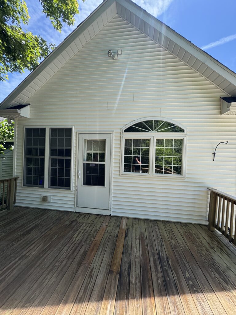 White house exterior with a wooden deck, vinyl siding, and a gable roof under a blue sky with trees nearby.