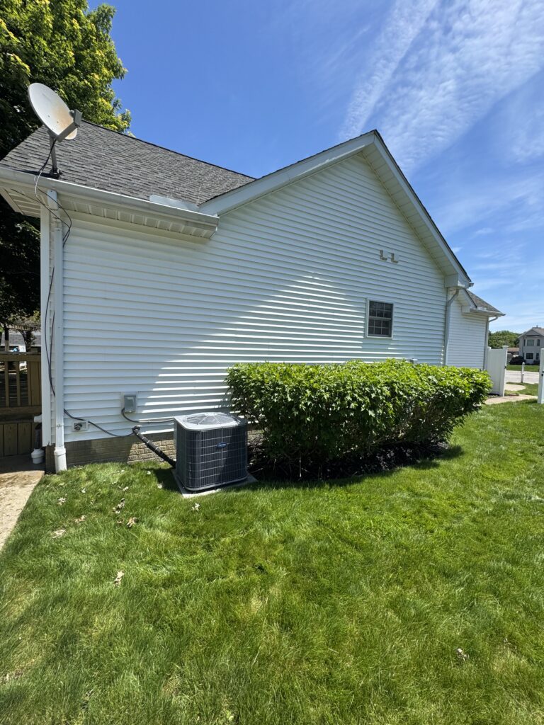 White siding house with a satellite dish on the roof, and a ground-mounted air conditioner unit beside a trimmed hedge under a blue sky.