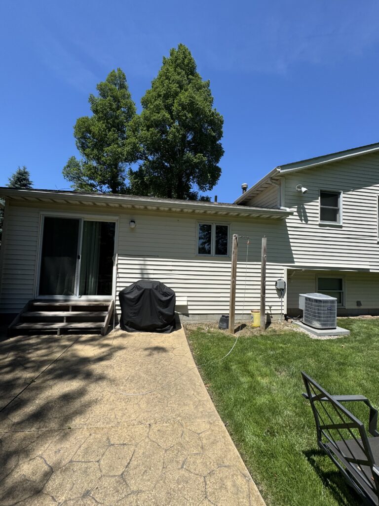 Backyard of a house with a large leafy tree, sliding door, and a covered grill on a concrete patio under a clear blue sky.