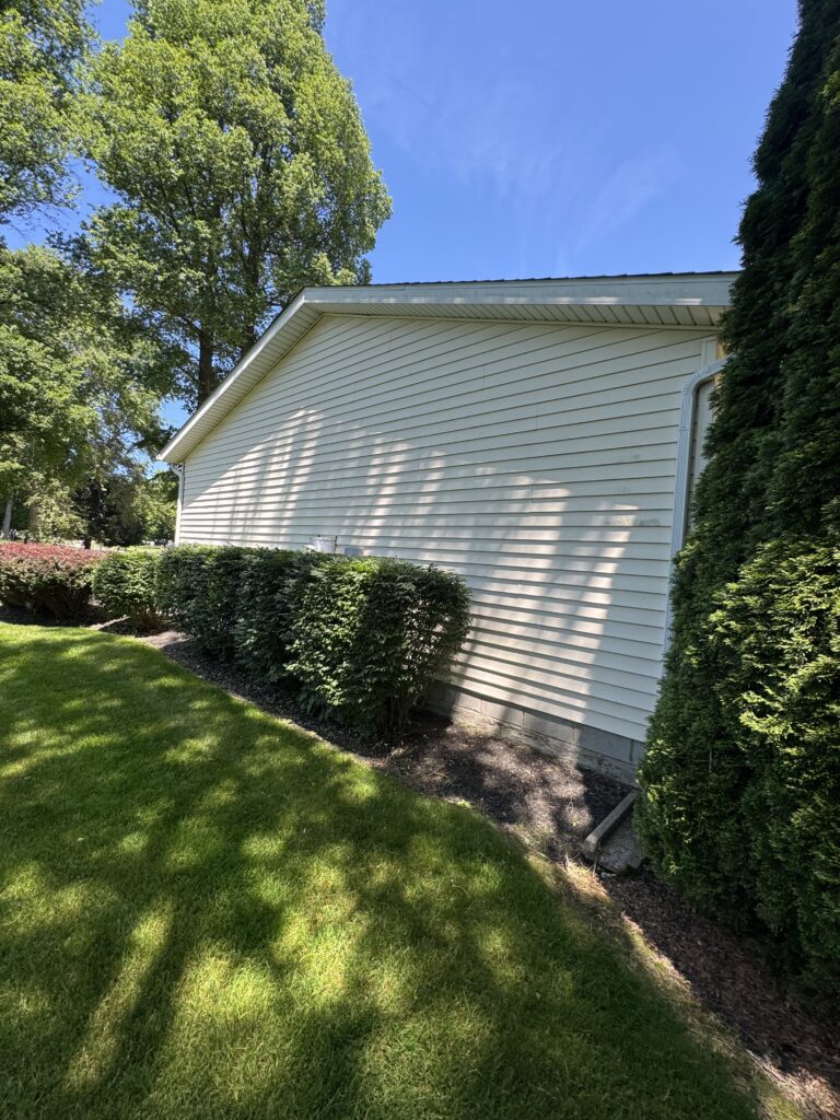 Side of a beige house with white gutter, bordered by green shrubs and tall trees under a bright blue sky.