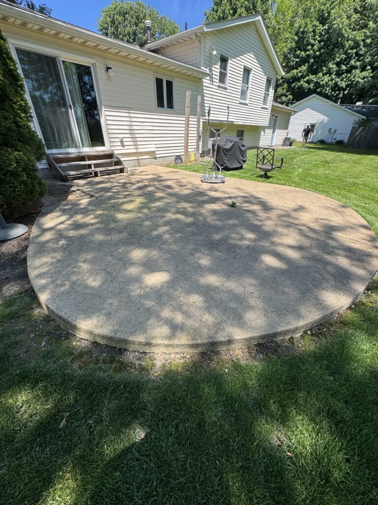 Backyard with a large circular concrete patio beside a beige siding house, sliding glass doors, and a grill on the patio edge. A grassy lawn surrounds the area.