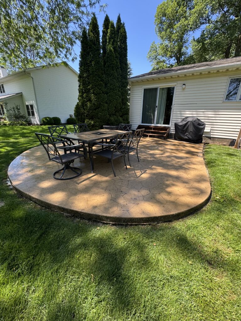 Backyard with a circular stamped concrete patio, outdoor dining table and chairs, a grill covered near the house, and tall evergreen trees in the background.