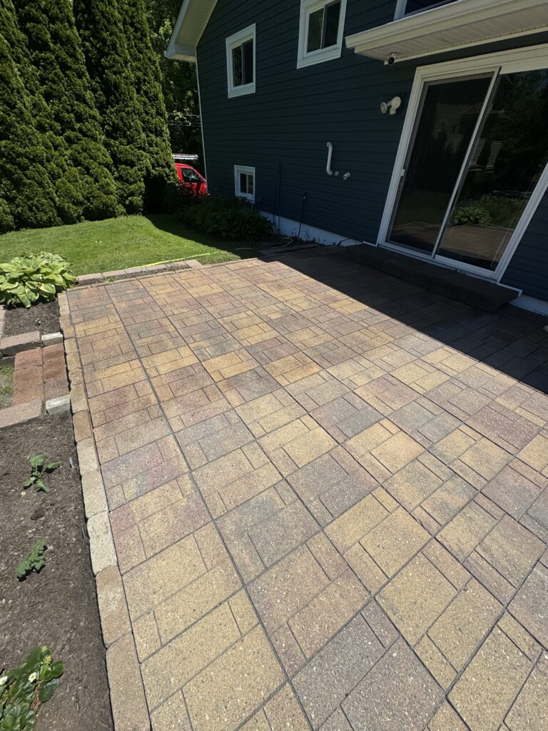 Backyard patio with tan-brown brick pavers beside a blue house, sliding glass door, and a tall evergreen hedge on the left.