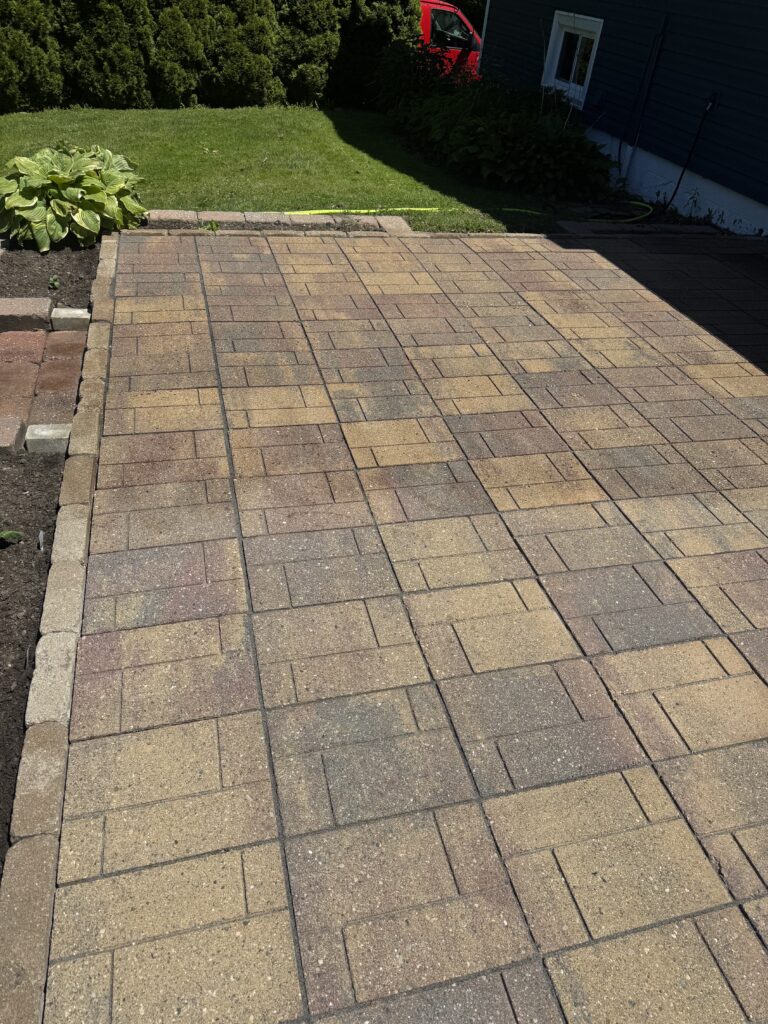 Brick patio extending across the image with a small edging wall, a green lawn behind, and shrubs on the left edge.