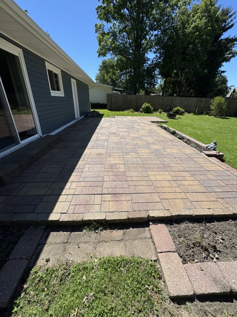 Backyard patio with tan brick pavers along a blue house, sliding glass door on left and a small window, wooden fence in the yard beyond.