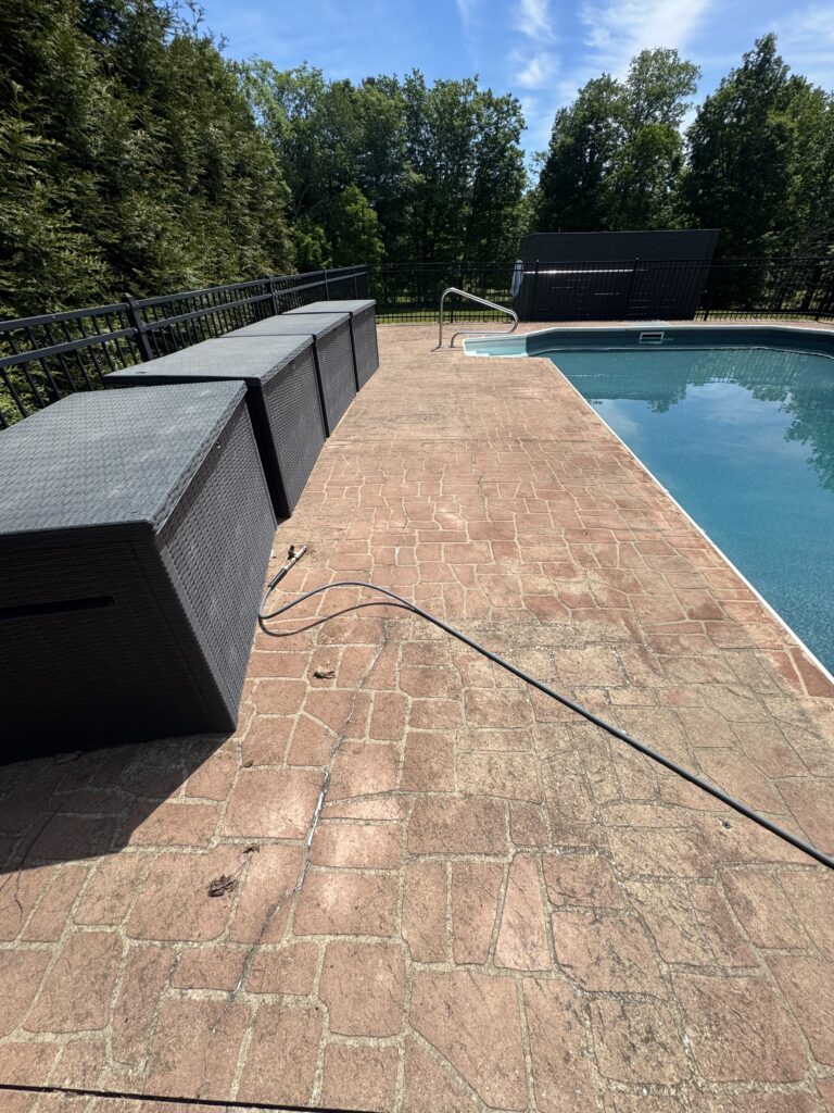Poolside brick patio with gray wicker storage benches along a black fence, next to a blue swimming pool and metal handrail, backed by trees and a clear sky.