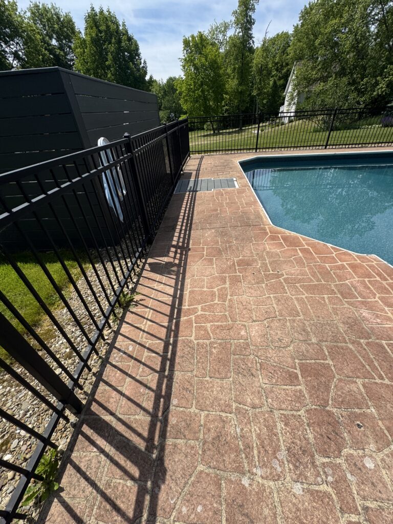 Backyard pool area with red brick paving, a black metal fence, and a dark privacy screen on the left; trees beyond the yard.