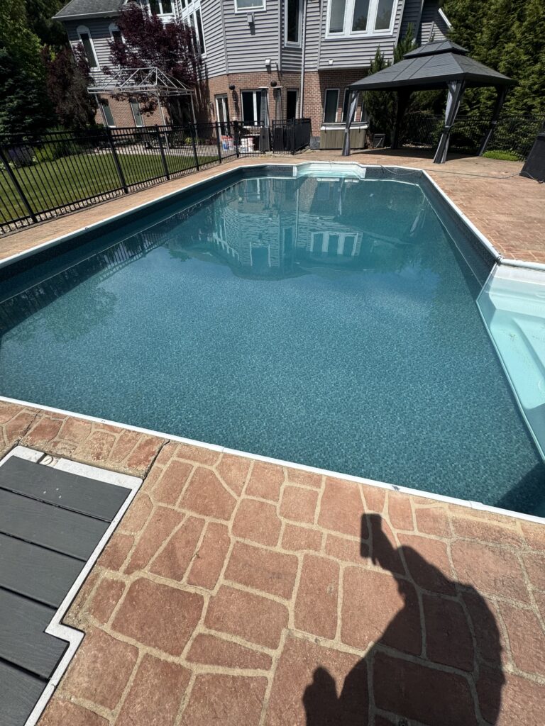 Backyard in-ground pool with brick-paver patio, black metal fence, and a gazebo near a gray-brick house; pool water reflects the building's image.