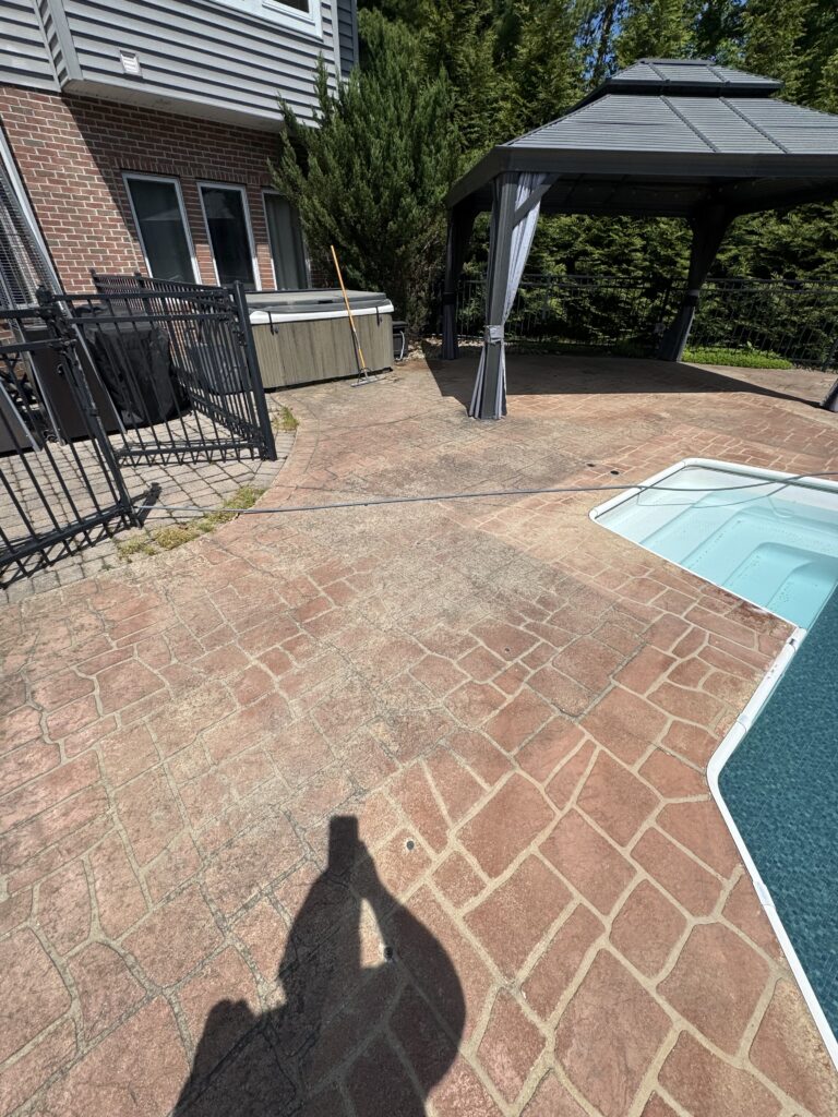 Backyard patio area with a brick house, hot tub, black metal fence, and a grey gazebo on a stamped concrete surface, sunny day.