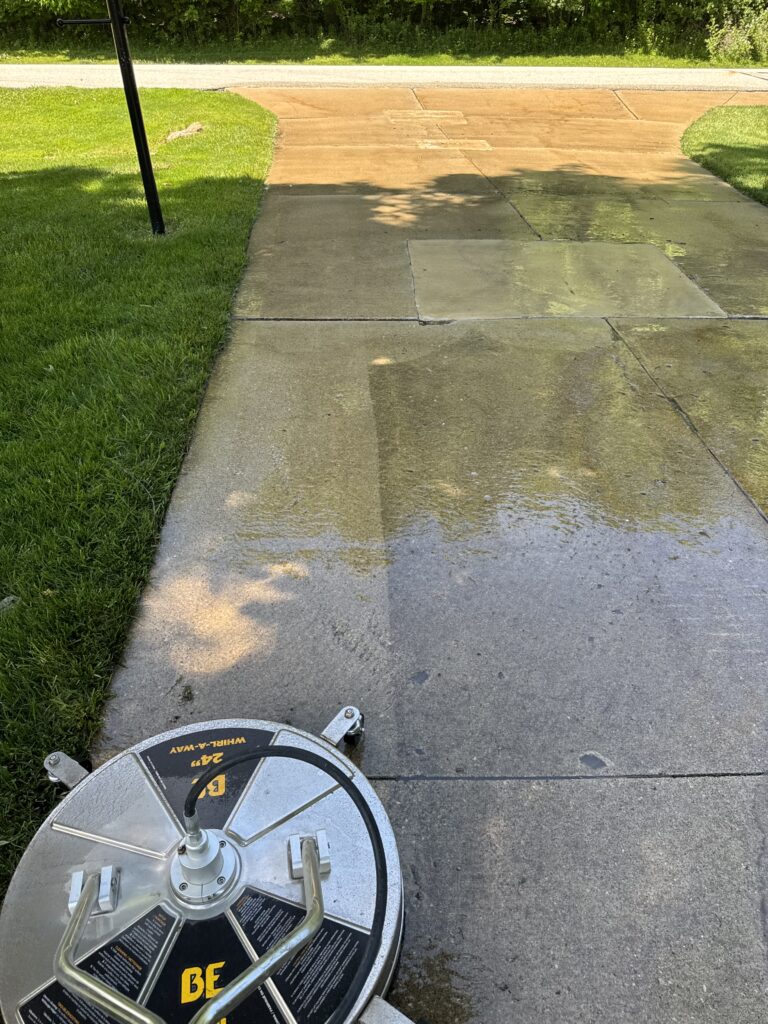 Partially visible lawn mower wheel in the lower left, on a wet concrete driveway with green grass on the left and a sunlit driveway ahead.