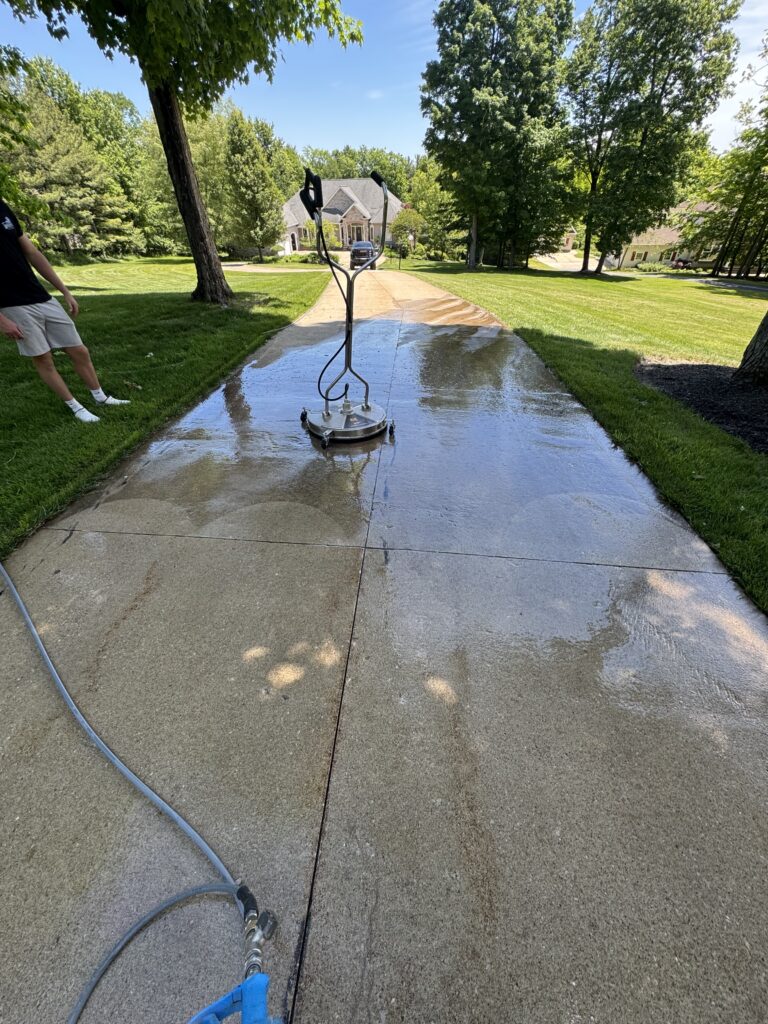 Wheeled pressure washer on a wet concrete walkway in a green suburban yard, with a person standing to the left and a house in the distance.