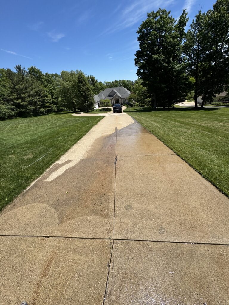 Long driveway leads to a suburban house with a dark pickup parked at the entrance, flanked by green lawns and trees under a blue sky.