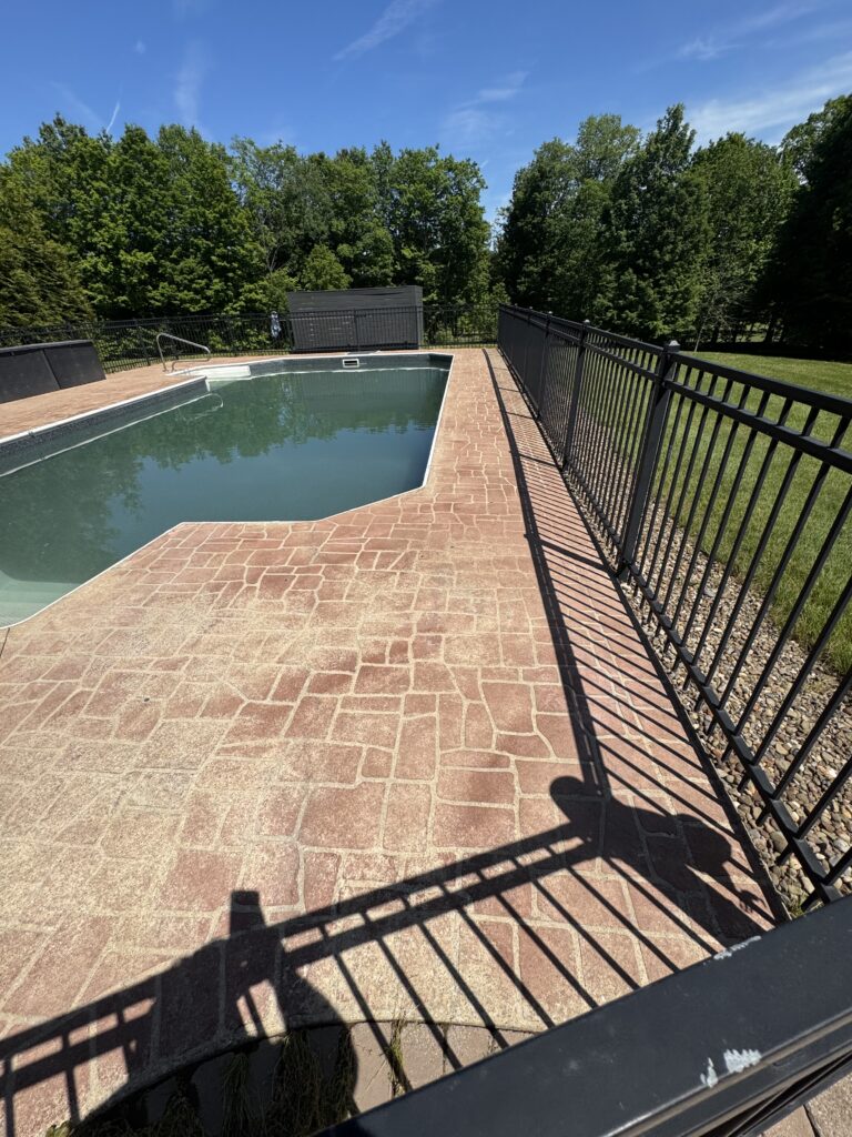 Outdoor pool surrounded by brick patio and a black metal fence, with green trees and a blue sky in the background.