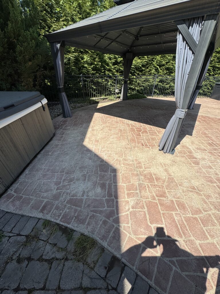 Patio area with a dark gray gazebo and curtains, brick-patterned pavers, and surrounding greenery along a metal railing fence.