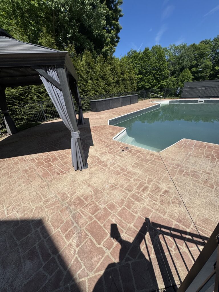 Outdoor pool area with a brick-paved deck, curtained cabana on the left, and tall green trees against a clear blue sky.