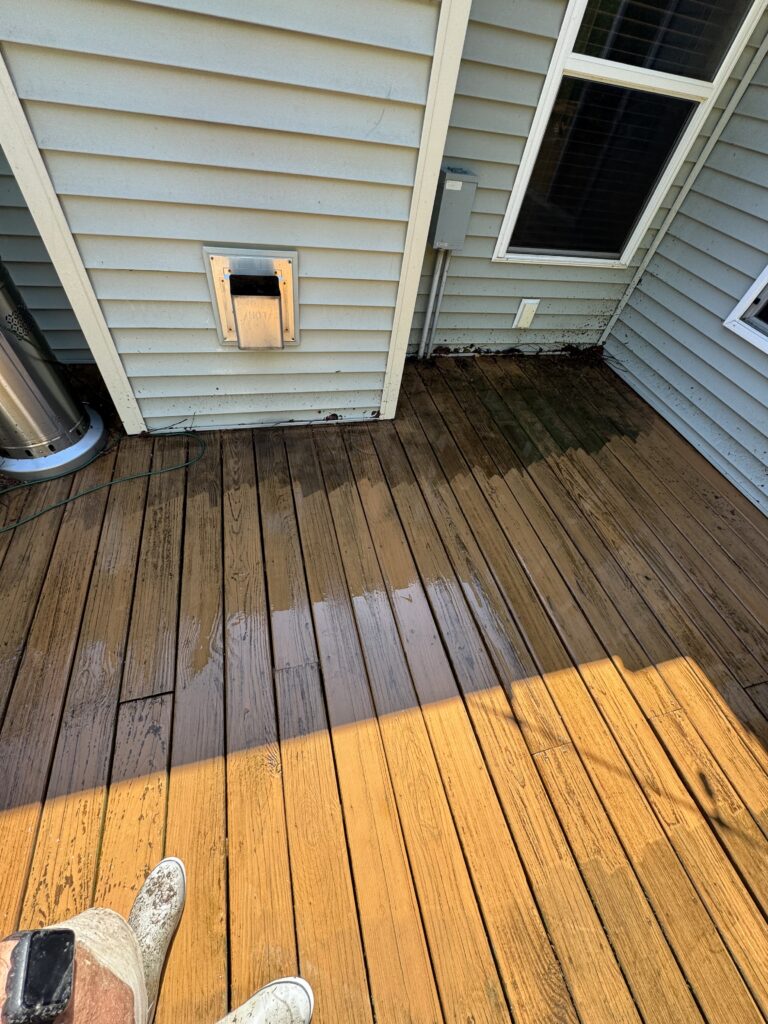 Photo from a person’s feet on a wet wooden deck, with beige siding, a vent box on the wall, and a window to the right; sunlight casts a yellow stripe across the boards.