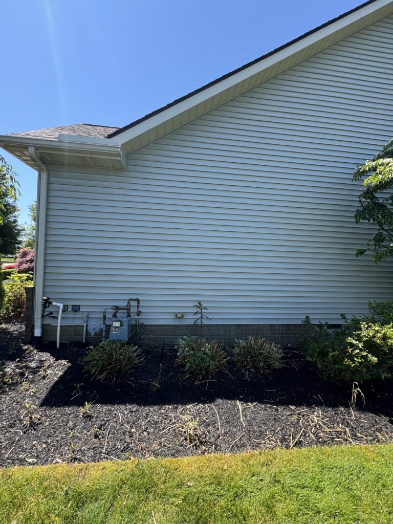 Side view of a blue house showing vinyl siding, gutter, and downspout with utility meters at the foundation near a mulched planting bed.