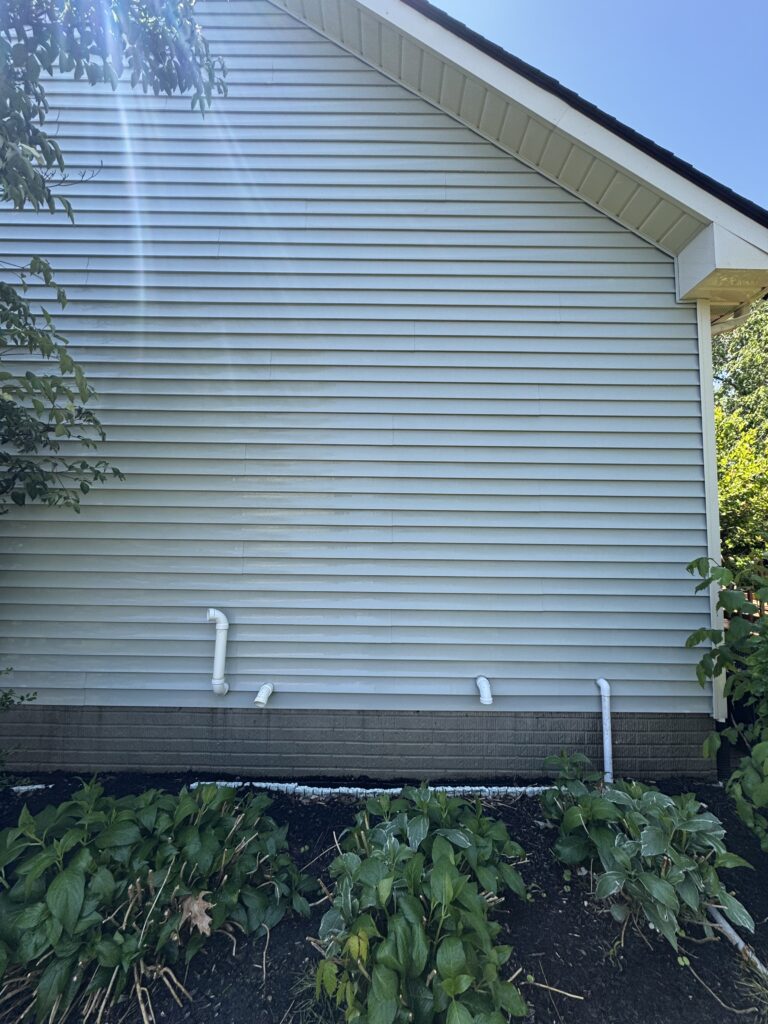 Side of a light blue house with horizontal siding, white trim along the roof, and multiple white downspouts along the foundation, with green bushes in the foreground.