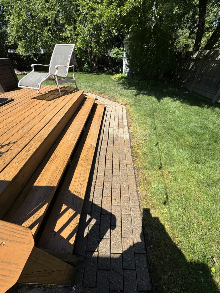 A gray outdoor chair on a wooden deck beside a brick patio, with green lawn and trees in the background.