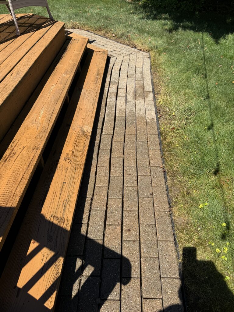 Wooden deck stairs with a chair leading to a curved brick walkway beside a green lawn in sunlight.