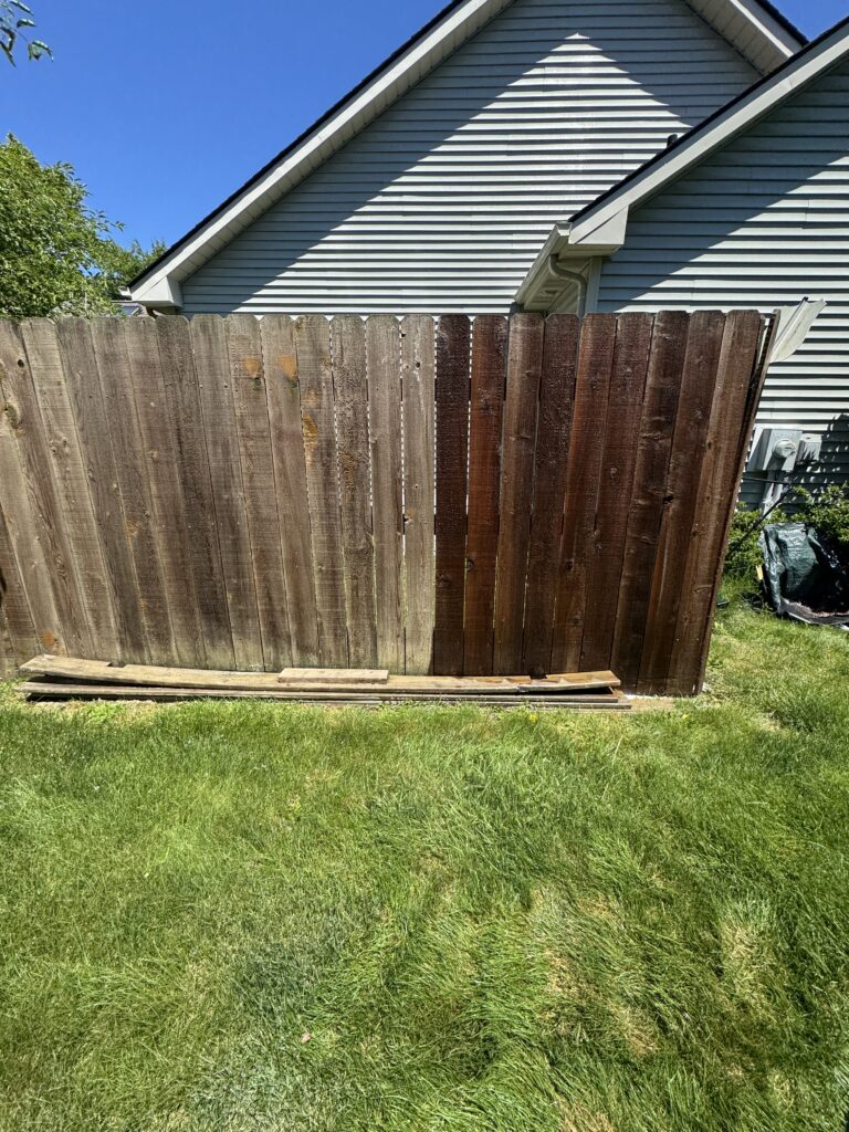 Tall weathered wooden privacy fence leaning inward in a backyard, with a blue house and clear sky behind it.