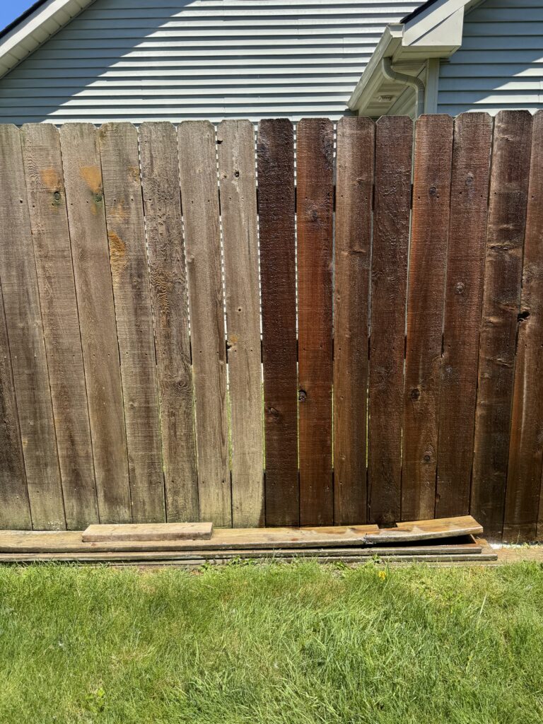 Brown weathered wooden privacy fence in front of a light-blue house, with a guttered roofline visible above and grass at the bottom.