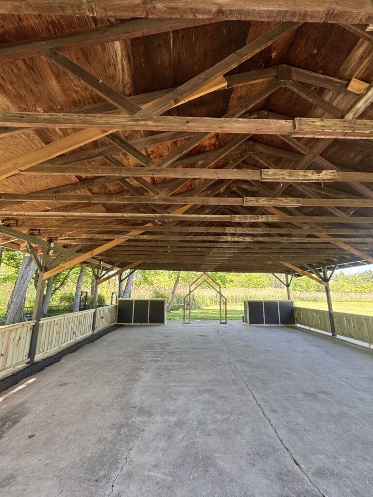 Open-air wooden pavilion with exposed trusses and a concrete floor, facing a grassy field with seating areas nearby.