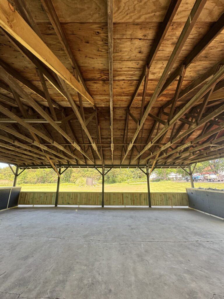 Open-sided pavilion with a wooden truss roof and concrete floor, looking toward a grassy field and parked trucks beyond.