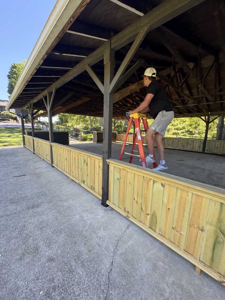 Man on a red ladder under a wooden shelter, hammering or adjusting the dark painted beams while wearing a light-colored cap.