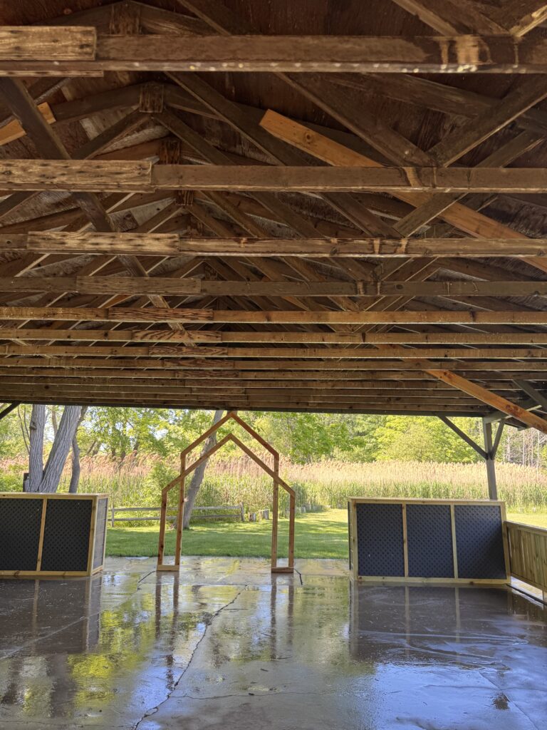 Open-air pavilion interior with exposed wooden roof trusses and a wet, reflective concrete floor; outdoor greenery visible beyond.
