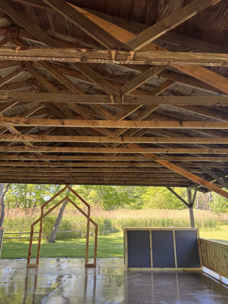Interior view of a wooden pavilion with exposed angled roof trusses, open to a grassy yard and trees beyond, on a wet concrete floor.