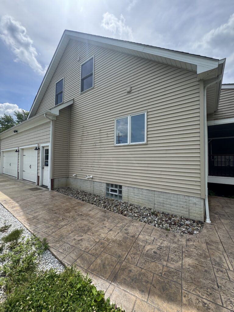 Two-story beige house with vinyl siding, gabled roof, and two first-floor windows; white garage doors along left side and decorative landscaping stones all Serviced in Madison, OH by Wiley's Pressure Washing
