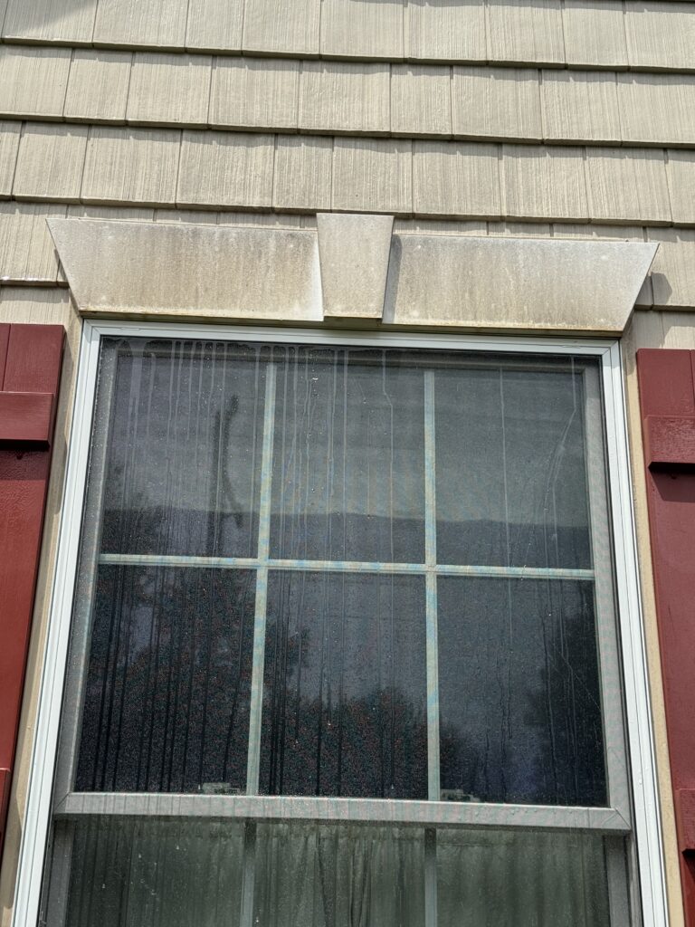 Exterior window with a white decorative awning/overhang above beige siding, rain streaks on the glass windowpane before cleaning from Wiley's Pressure Washing Services in Madison, OH