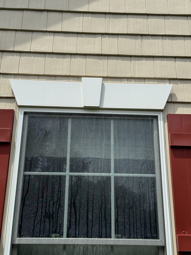 Exterior window with a white decorative awning/overhang above beige siding, rain streaks on the glass windowpane after cleaning from Wiley's Pressure Washing Services in Madison, OH