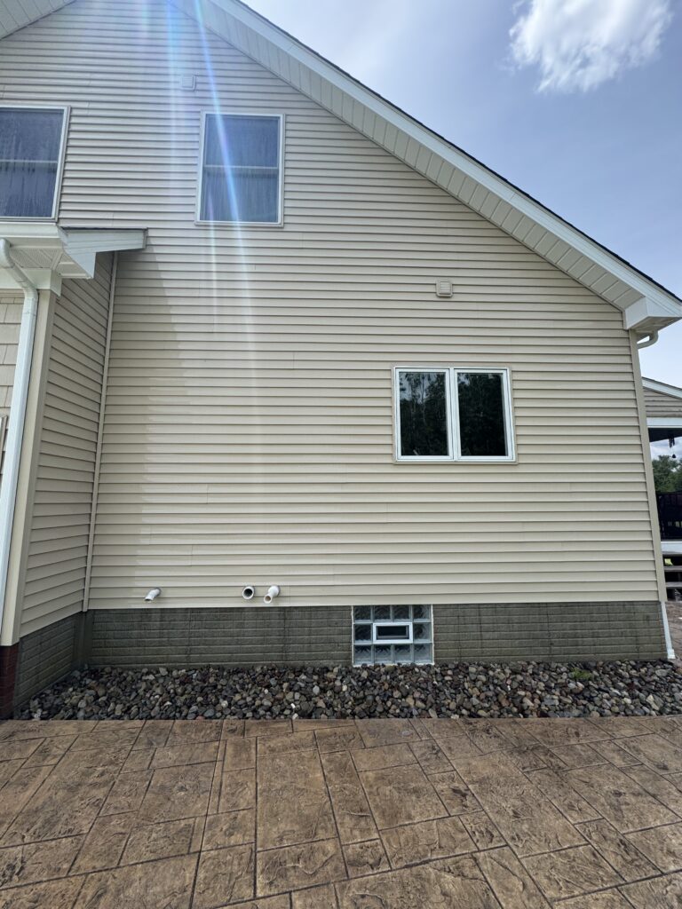 Backyard view of a beige vinyl-sided house with two upstairs windows and a sun flare across the wall, gravel at the foundation, and a patterned concrete patio in the foreground all cleaned in Madison, OH by Wiley's Pressure Washing Services 