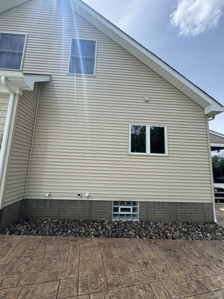 Backyard view of a beige vinyl-sided house with two upstairs windows and a sun flare across the wall, gravel at the foundation, and a patterned concrete patio in the foreground all cleaned in Madison, OH by Wiley's Pressure Washing Services 