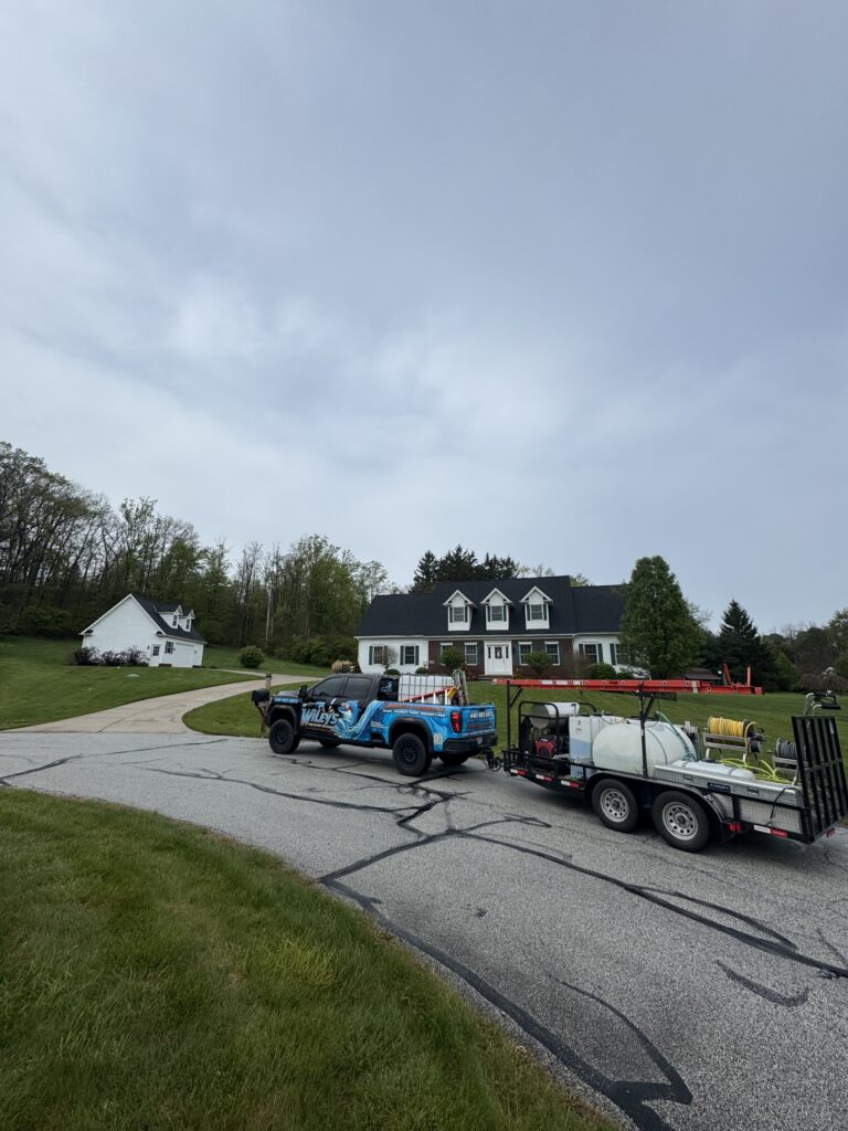 Blue service pickup and trailer with hoses and tanks parked in a suburban driveway in front of a two-story house with a dark roof and white siding.