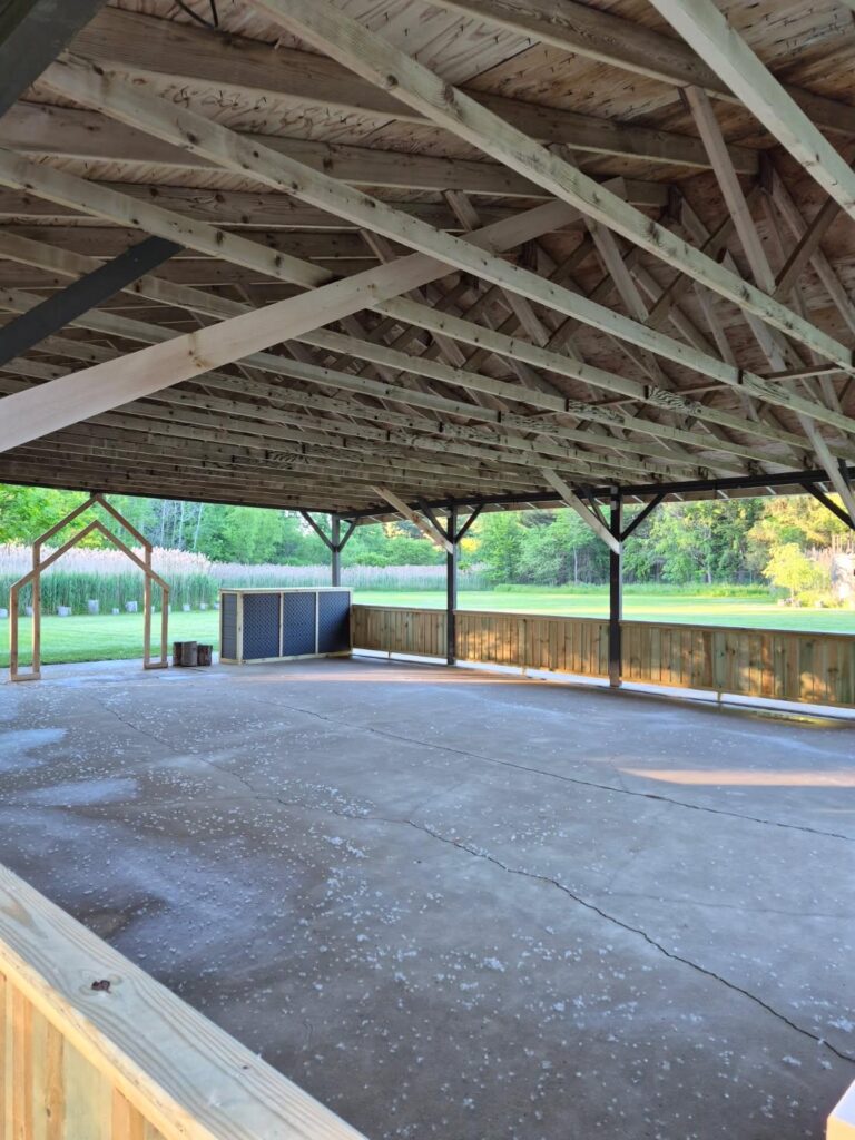 Open-air wooden pavilion with exposed roof trusses and a concrete floor, set in a grassy park with trees in the background; there is a blue cabinet and wooden railing along the edge.