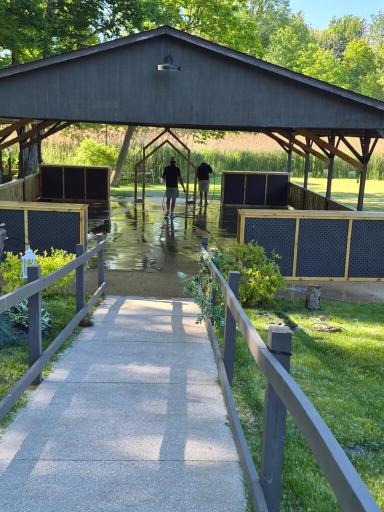 Two men hose down a wet concrete floor inside a dark wooden pavilion with lattice walls and surrounding greenery.