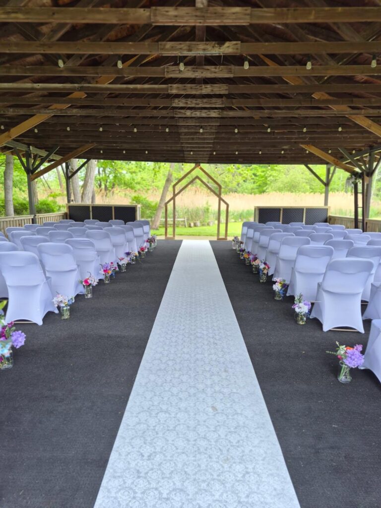 Outdoor wedding ceremony setup under a wooden pavilion, with white chairs lining a light-blue patterned aisle toward a wooden arch all set up after services from Wiley's pressure Washing in Madison, OH